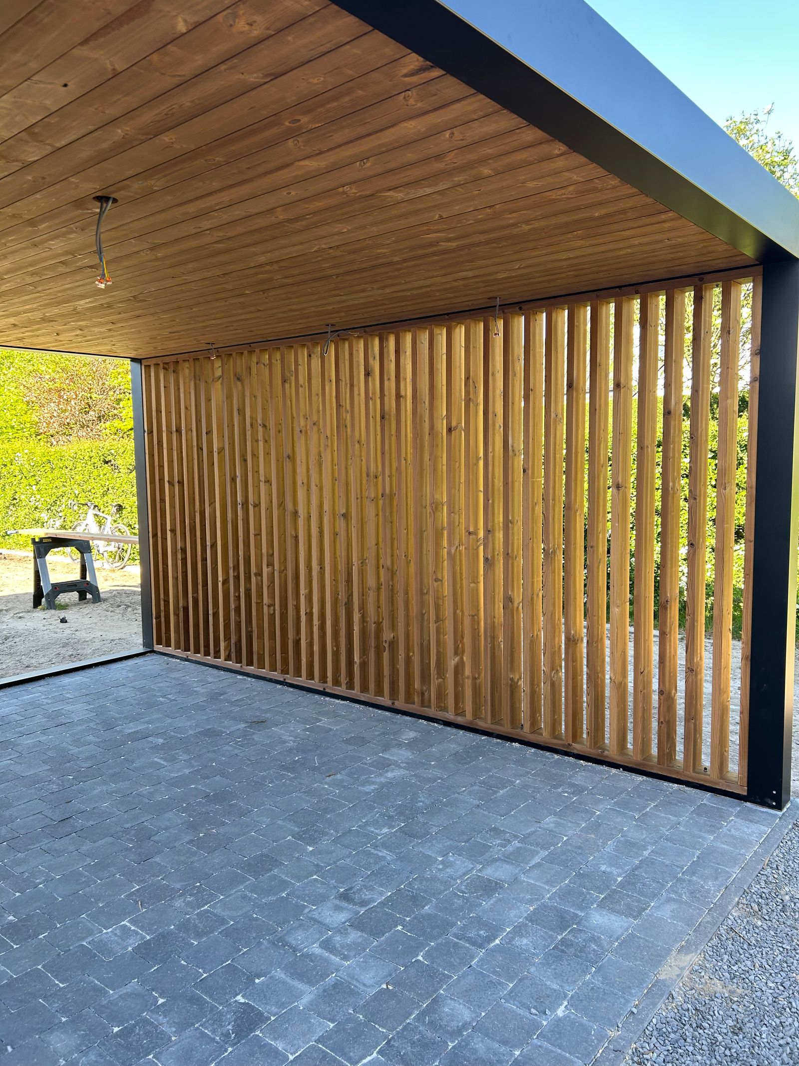 Het houten plafond geeft een warme sfeer, terwijl de open latstructuur aan de zijkant zorgt voor een mooie balans tussen privacy, lichtinval en ventilatie.